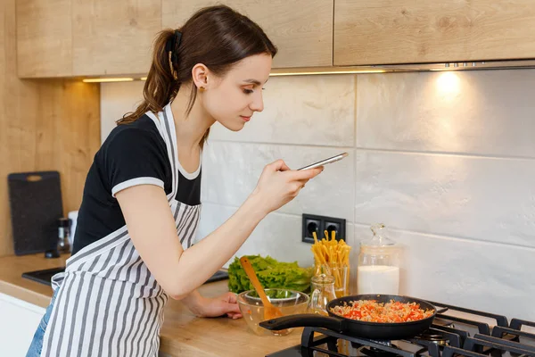 Young woman blogger in apron taking photo with phone while cooking food at kitchen. Preparing meal with frying pan on gas stove. Concept of domestic lifestyle, housewife leisure and culinary blogging — Stock Photo, Image