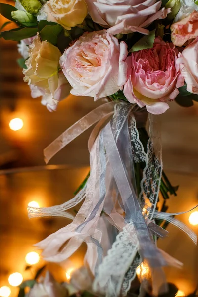 Delicate and very beautiful wedding bouquet of roses, greenery, eustoma standing on mirror table. Bridal trendy flowers in pastel colors on the background of the evening lighting bulbs. — Stock Photo, Image