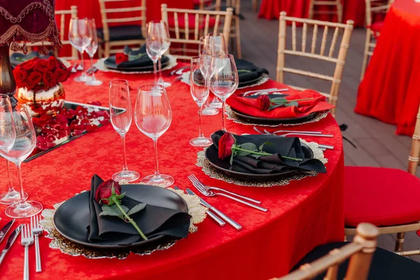 客人餐桌设置为黑色,红色和金色风格的宴会.精致的晚餐：装饰、桌布、餐巾纸盘、新鲜玫瑰、眼镜、餐具。屋顶上的派对庆祝活动，户外. — 图库照片