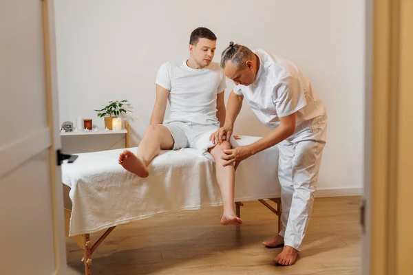 Bellissimo massaggiatore in uniforme bianca che fa terapia manuale per il ginocchio di un giovane atleta. Massaggio professionale e riabilitazione per sportivi. Concetto di benessere, corpo e assistenza sanitaria — Foto Stock