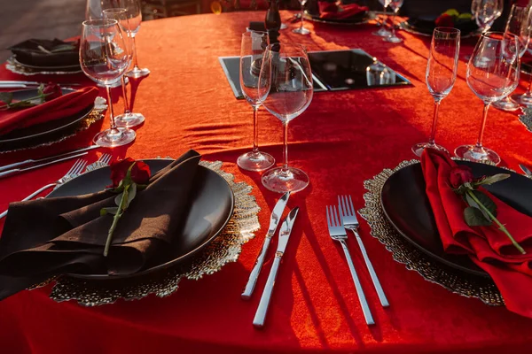 Guests table setting for banquet in black, red and gold style. Elegant dinner: decor, tablecloth, plates with napkins and fresh roses, glasses, cutlery. Themed party celebration on the roof, outdoor.
