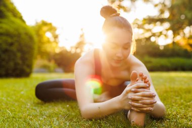 Young smiling woman doing stretching exercise in the park it sunset rays. Selective focus on feet. Concept of outdoors yoga, fitness, active and sporty lifestyle. Flexible girl in black sportswear.