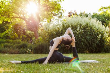 Young woman in black clothing practice yoga on mat in the summer park. Slim girl in sunset rays doing stretching exercise. Concept of outdoors yoga, fitness, active and sporty lifestyle.