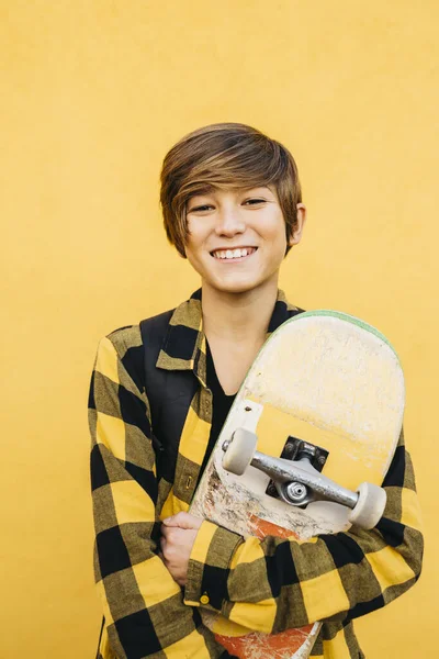 Portrait of a smiling teenager student boy with a skateboard in front of a yellow background