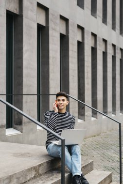Genç adam, merdivenlerde oturuyor, dizüstü bilgisayar kullanıyor ve kırmızı bir akıllı telefondan konuşuyor.