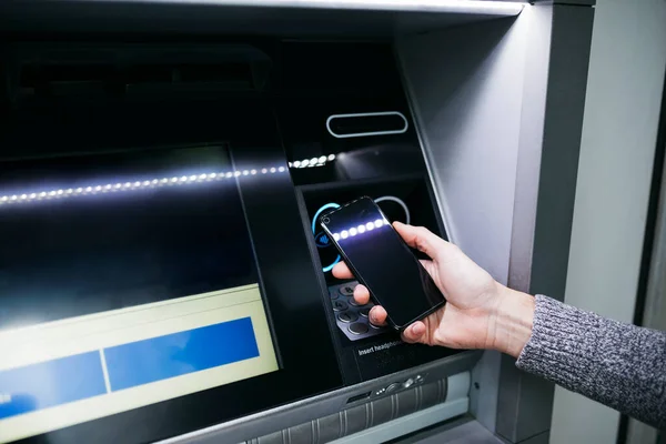 Man`s hand with an smartphone using an ATM street machine to withdraw some cash