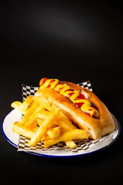 Frankfurt sausage with ketchup and mustard accompanied with french fries on a white plate over a black background