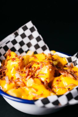 Fried potatoes with melted cheese and topped with pieces of bacon on a white plate over a black background