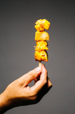Human hand holding a brochette of fried potatoes with melted cheese and topped with bacon over a grey background