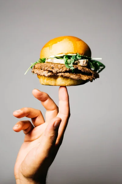 Human hand holding with his finger a cheeseburger with old dry cheese and lettuce, over a grey background