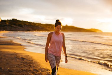 Spor kıyafetli genç bir kadın gün doğumunda sahilde yürüyor.