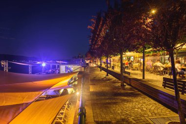 Arona, İtalya. Corso Guglielmo Marconi 'nin gece manzarasına bakın. İnsanlar yürüyor ve diğerleri şehrin göl kıyısındaki restoranların açık hava masalarında oturuyor. 10 Haziran 2022.
