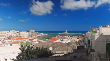 Vieste, İtalya. Açık gökyüzü ve beyaz bulutlarla dolu bir yaz günü Vieste manzarası. Uzakta deniz feneri ve Colombo gezinti güvertesinin küçük plajı. Gerçek zamanlı video. 5 Eylül 2022.