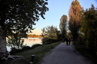 Sonbaharda gün batımında nehir kenarındaki bir parkta gezinen insanlarla dolu çakıl yolu.