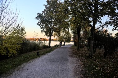 Sonbaharda gün batımında nehir kenarındaki bir parkta gezinen insanlarla dolu çakıl yolu.