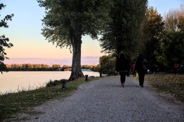 Sonbaharda gün batımında nehir kenarındaki bir parkta gezinen insanlarla dolu çakıl yolu.