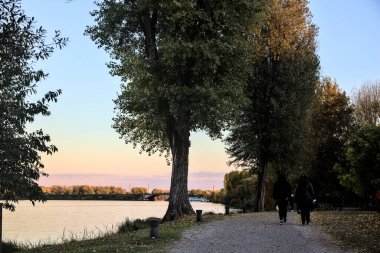 Sonbaharda gün batımında nehir kenarındaki bir parkta gezinen insanlarla dolu çakıl yolu.