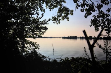 Gün batımında gölde ağaçların siluetiyle çerçevelenmiş.