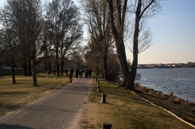 Gölün kıyısındaki bir parkta güneşli bir günde gezinen insanlarla dolu bir yol.