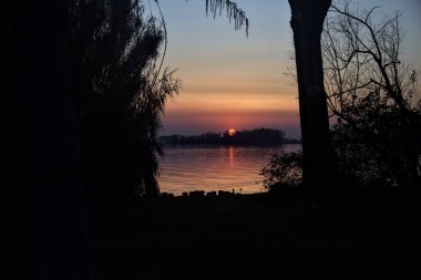 Uzaklardan görünen ağaçlarla çerçevelenmiş bir gölün üzerinde batan güneş.