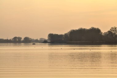 Gün batımında göl, uzakta bir orman ve suya düşen bir gökyüzü.