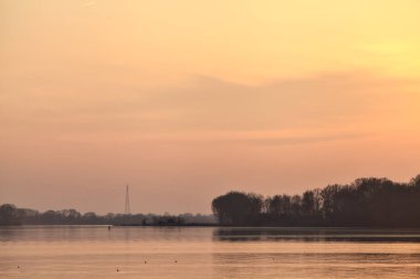 Gün batımında göl, uzakta bir orman ve suya düşen bir gökyüzü.
