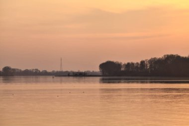 Gün batımında göl, uzakta bir orman ve suya düşen bir gökyüzü.