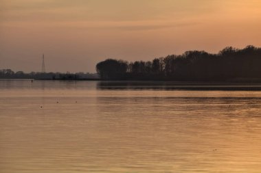 Gün batımında göl, uzakta bir orman ve suya düşen bir gökyüzü.