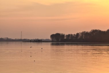 Gün batımında göl, uzakta bir orman ve suya düşen bir gökyüzü.