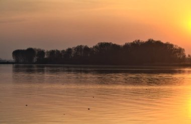 Gün batımında göl, uzakta bir orman ve suya düşen bir gökyüzü.