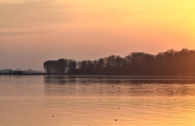 Gün batımında göl, uzakta bir orman ve suya düşen bir gökyüzü.