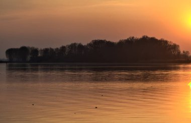 Gün batımında göl, uzakta bir orman ve suya düşen bir gökyüzü.