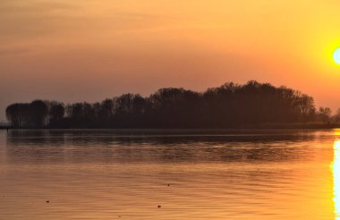 Gün batımında göl, uzakta bir orman ve suya düşen bir gökyüzü.