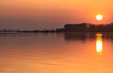 Gün batımında göl, uzakta bir orman ve suya düşen bir gökyüzü.