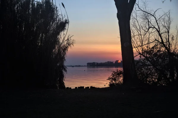 Uzaklardan görünen ağaçlarla çerçevelenmiş bir gölün üzerinde batan güneş.