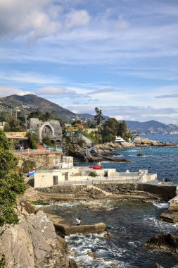 Güneşli bir günde deniz kenarında tarihi evler ve ağaçlarla dolu bir uçurum ve uzaktaki denize kadar uzanan bir kara şeridi.