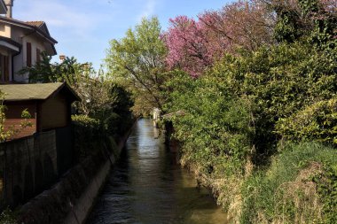 İtalya kırsalında güneşli bir günde suya yansıyan evler ve ağaçlarla çevrili su akıntısı.