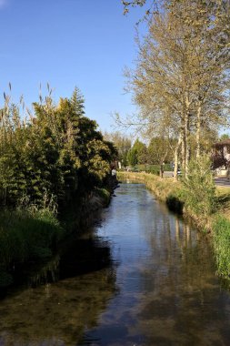 İtalya kırsalında güneşli bir günde suya yansıyan evler ve ağaçlarla çevrili su akıntısı.