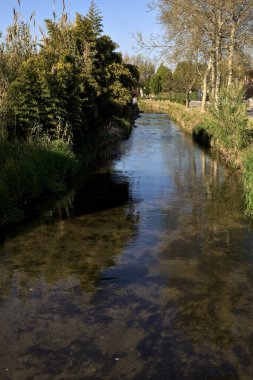 İtalya kırsalında güneşli bir günde suya yansıyan evler ve ağaçlarla çevrili su akıntısı.