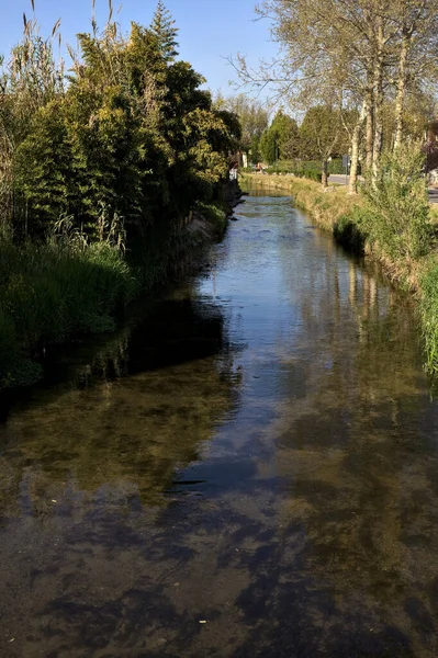 İtalya kırsalında güneşli bir günde suya yansıyan evler ve ağaçlarla çevrili su akıntısı.