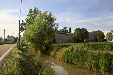 Güneşli bir günde uzakta bir kır evi olan bir dere ve ağaçlarla çevrili köy yolu.