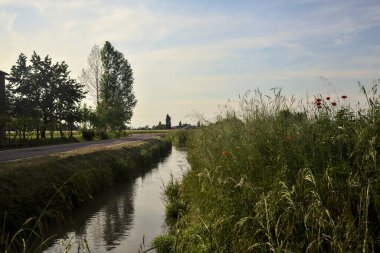 Güneşli bir günde uzakta bir kır evi olan bir dere ve ağaçlarla çevrili köy yolu.