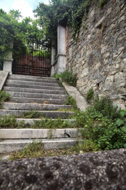 Bulutlu bir günde taş bir duvarın yanındaki paslı kapıya çıkan aşınmış taş merdiven.