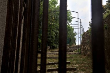 Ağaçlarla çevrili bir tepede merdiven ve ızgaranın içinden görünen taş bir duvar.