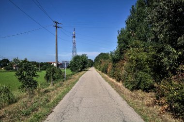 Gravel road bordered by fields with electricity poles at its edge and a country house in the distance on a summer day in the italian countryside