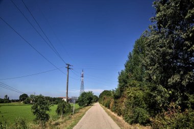 Gravel road bordered by fields with electricity poles at its edge and a country house in the distance on a summer day in the italian countryside