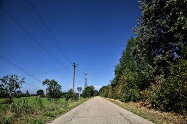 Gravel road bordered by fields with electricity poles at its edge and a country house in the distance on a summer day in the italian countryside