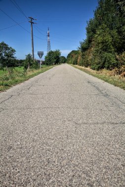 Gravel road bordered by fields with electricity poles at its edge and a country house in the distance on a summer day in the italian countryside