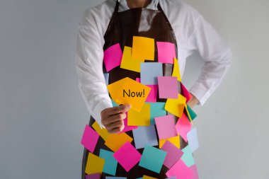 closeup woman wears apron with colorful sticky note paper holding a note with now word on one hand, concept and idea showing busy 
