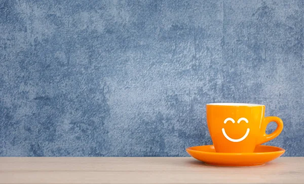 smiling face on orange cup of hot coffee isolated on wood table with blue background, happy morning